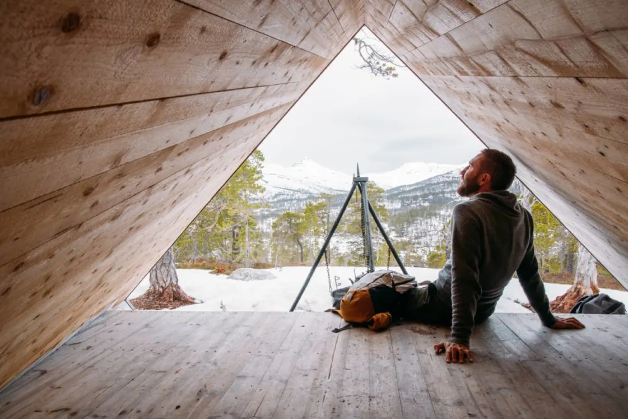 Man seated at cabin's edge, admiring snowy mountains